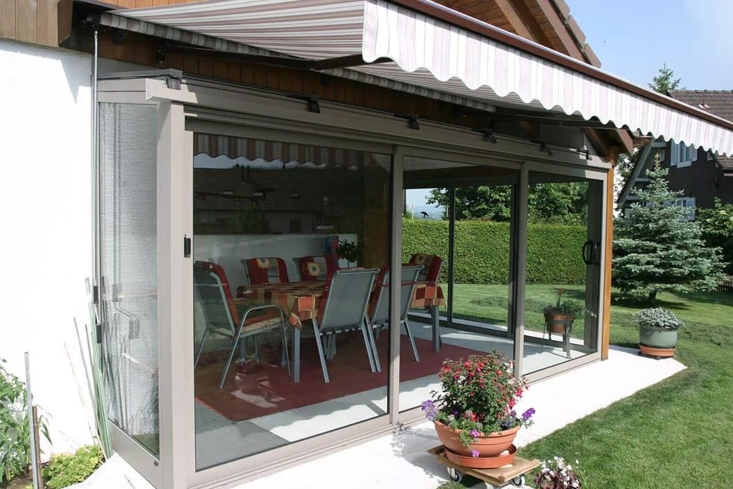 Beige aluminium schuifpui onder een overkapping met uitgerold knikarmscherm, met een eethoek erachter