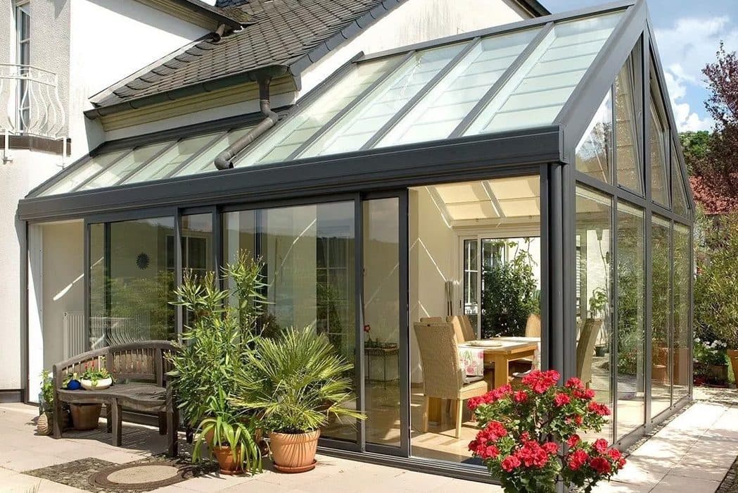Antraciet serre met glazen zadeldak en schuifpuien, met een tuinbank en bloeiende planten ervoor