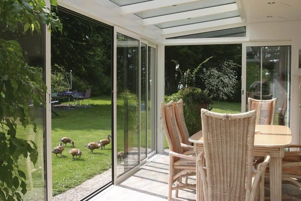 Tuinkamer met eettafel en rieten stoelen, met een open aluminium schuifpui naar het gazon