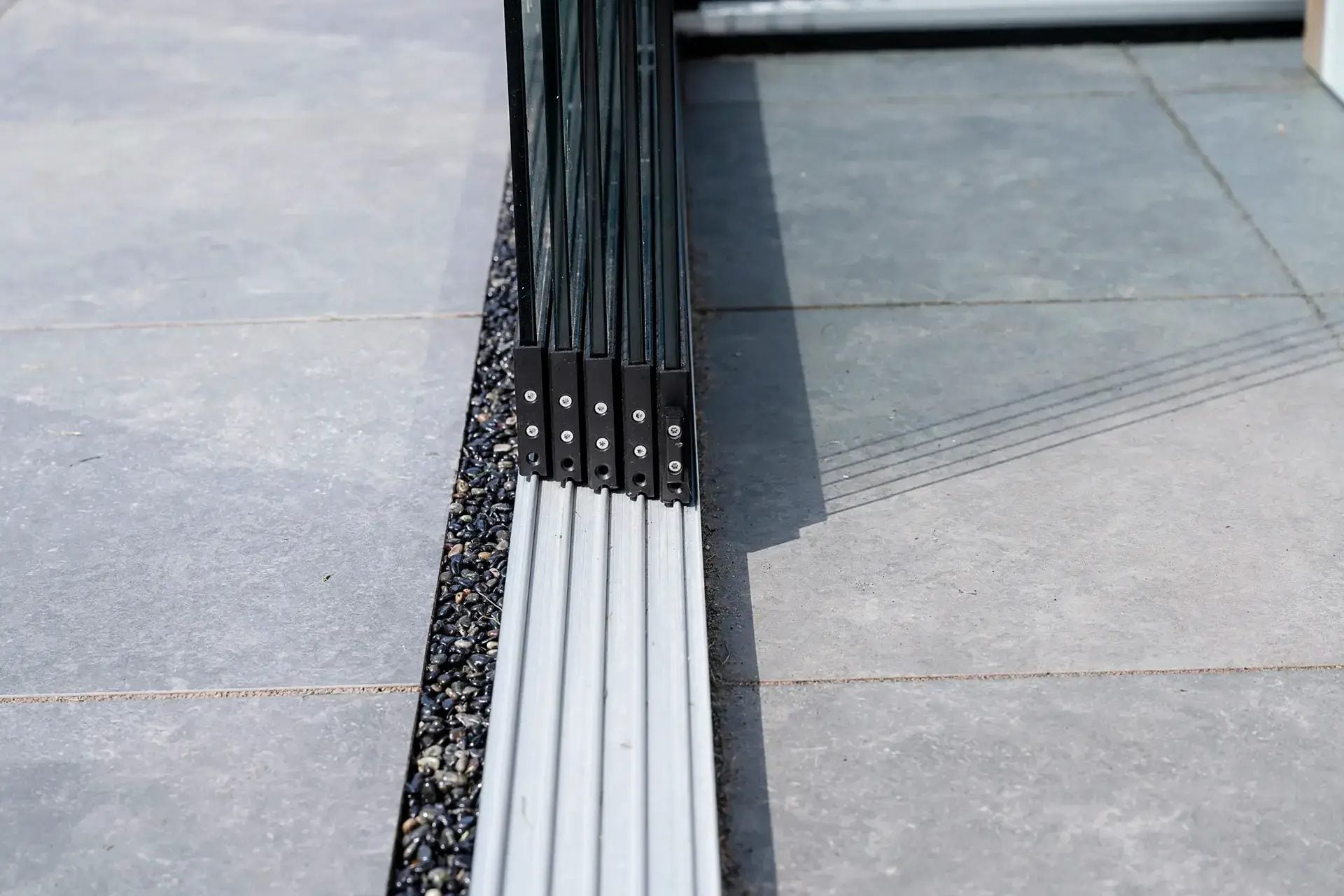 Close-up van de onderrail van een glazen schuifwand met de panelen naast elkaar geschoven, ingelaten in een tegelterras