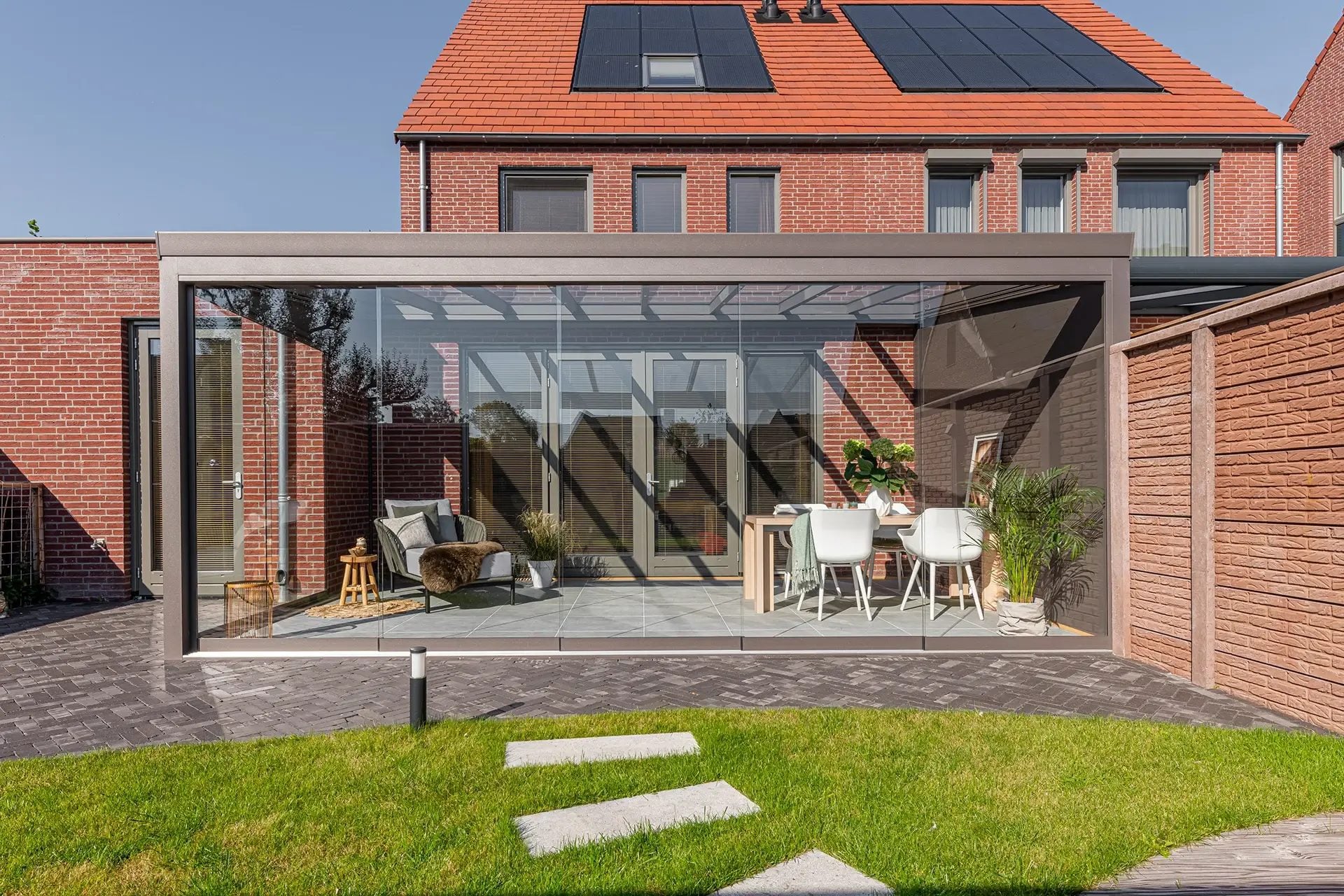 Bronskleurige veranda met glazen schuifwanden rondom achter een bakstenen woning, met loungestoel en eettafel