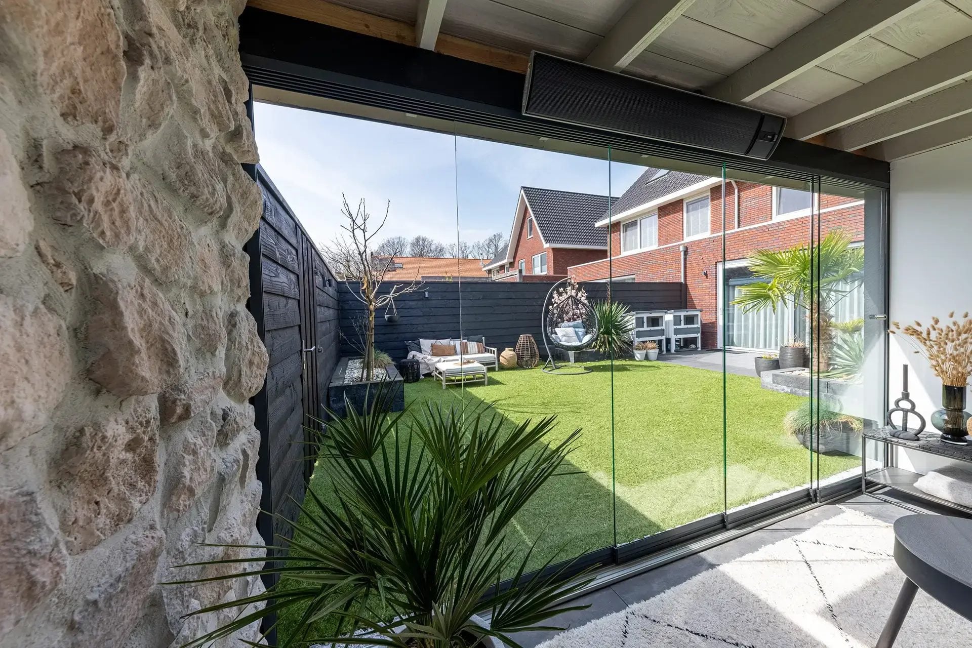 Glazen schuifwanden onder een houten overkapping met een natuurstenen muur, met zicht op de tuin