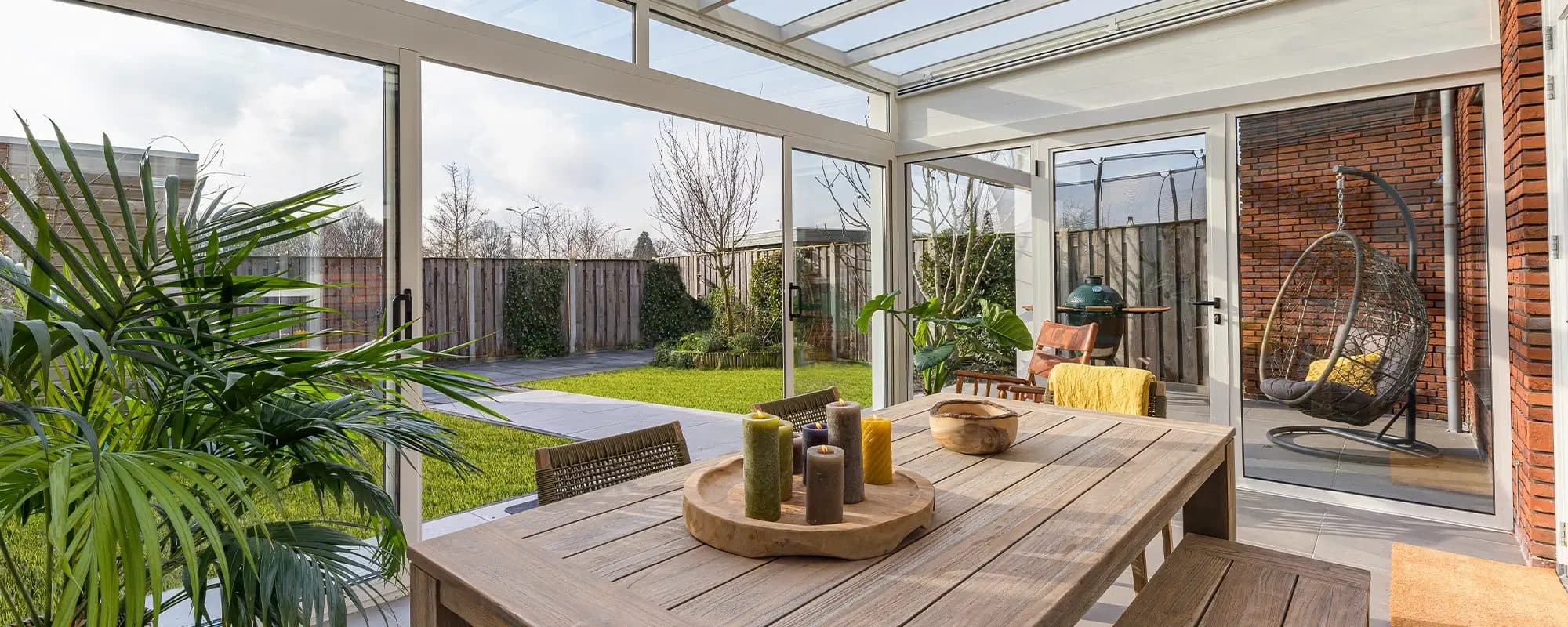 Houten eettafel in een witte tuinkamer met aluminium schuifpui en glasdak, met zicht op de tuin