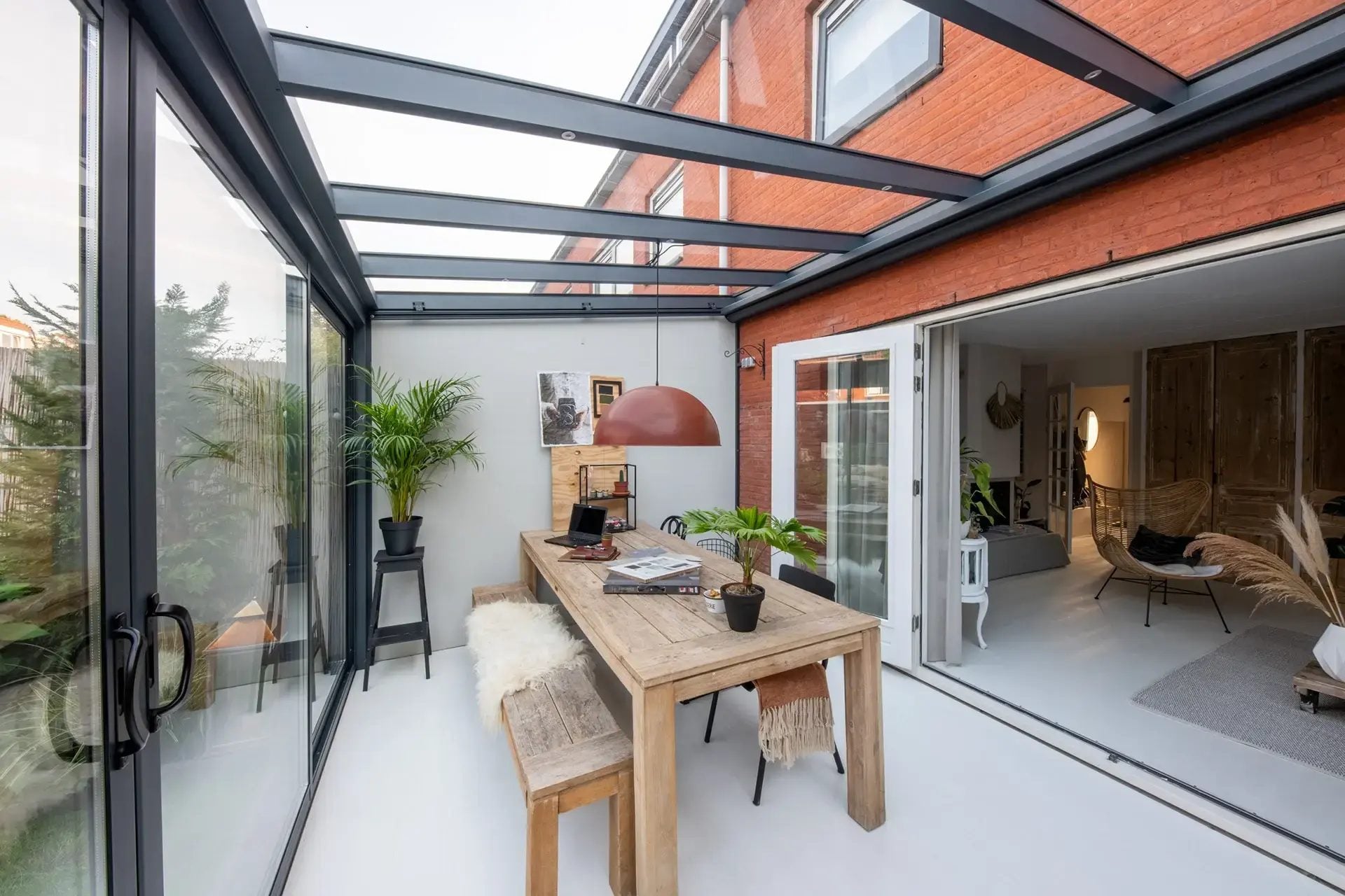 Lange eettafel in een tuinkamer met glasdak en aluminium schuifpui, met open deuren naar de woonkamer