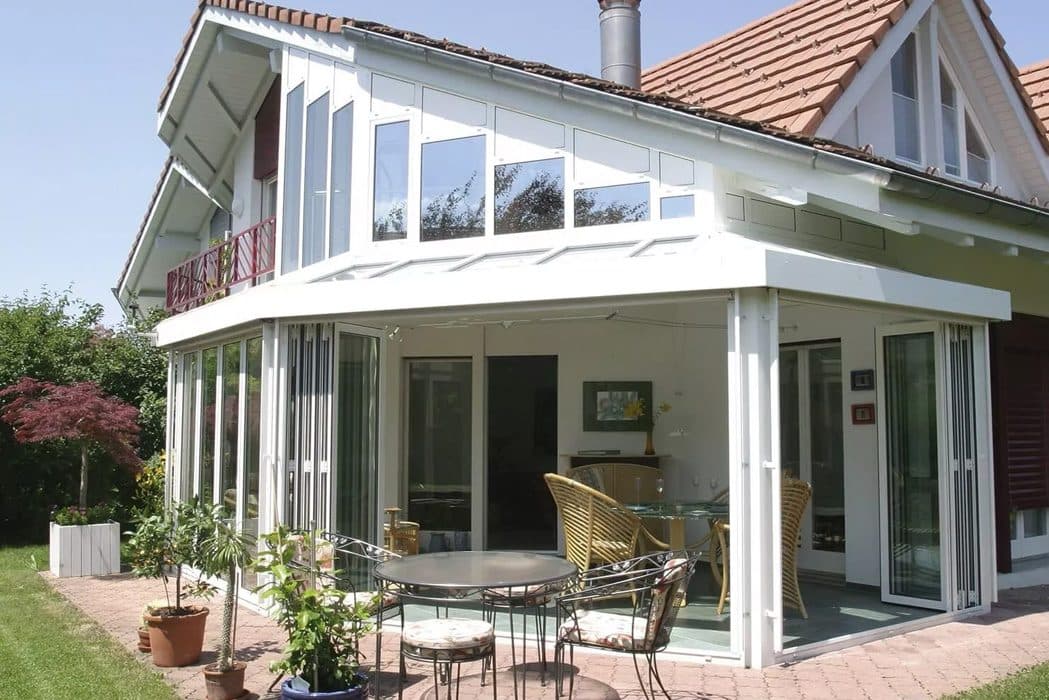 Witte serre met vouwwanden tegen een woning met puntdak, met een tuinset op een klinkerterras