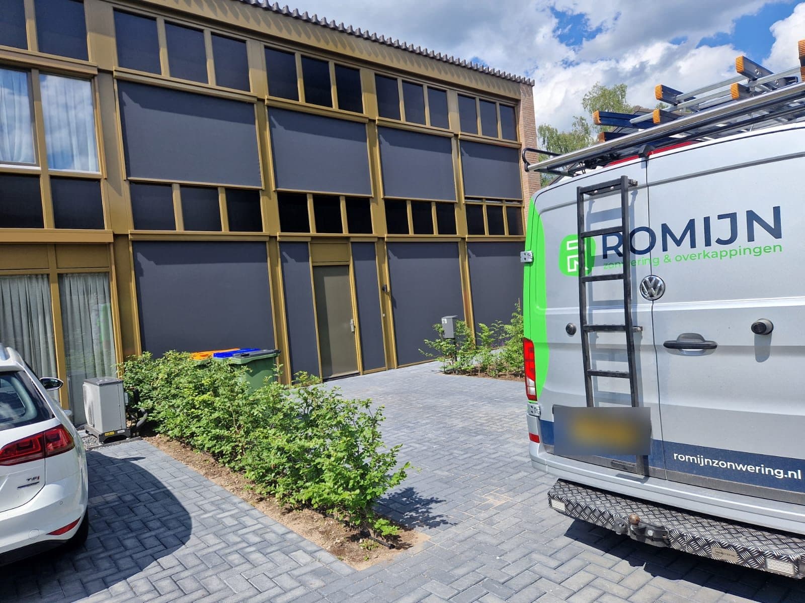 Bedrijfsbus van Romijn met ladders op het dak bij een gebouw met neergelaten screens voor de ramen