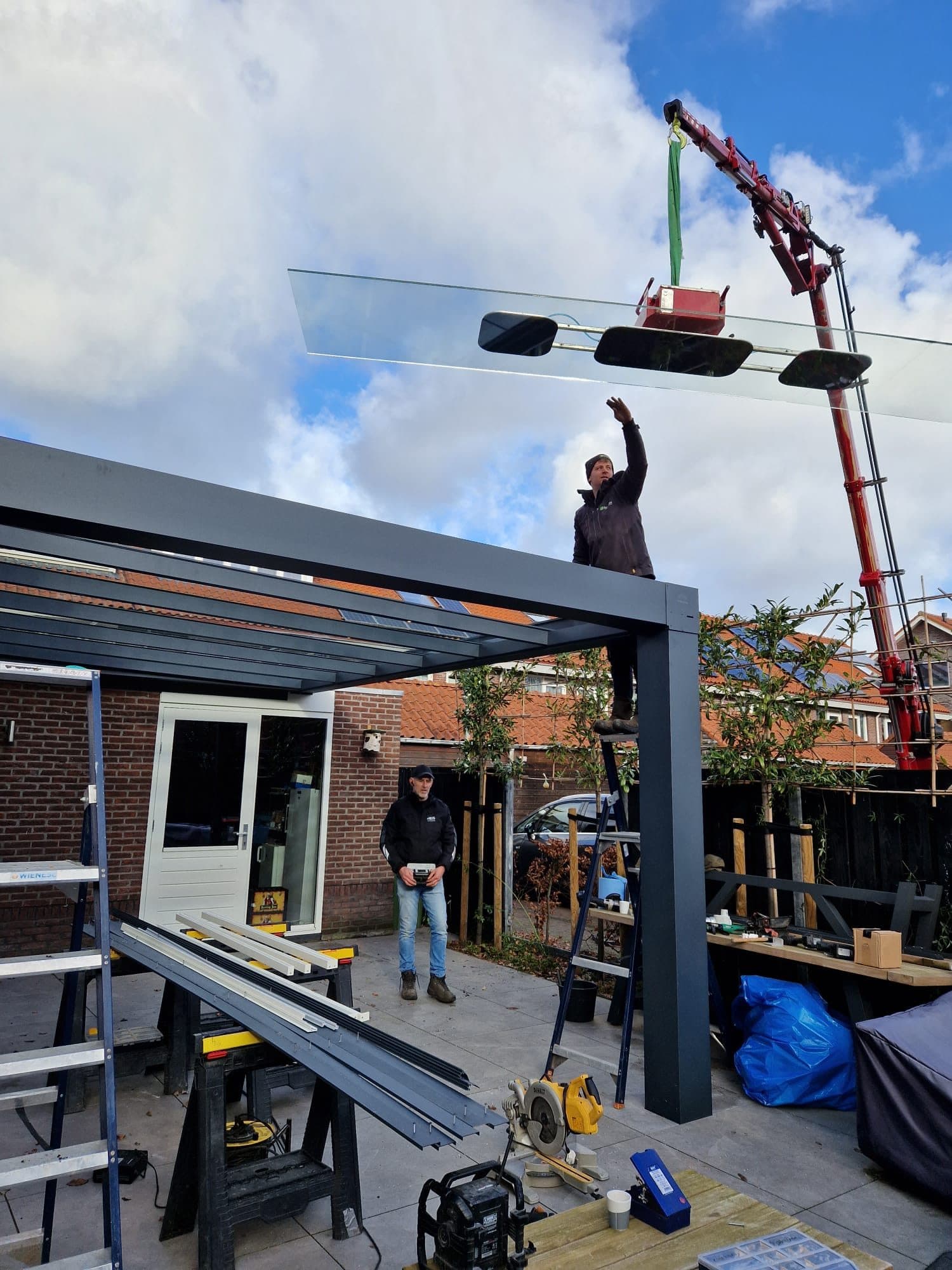 Monteur op het frame van een veranda leidt een glasplaat aan een kraan naar zijn plek, terwijl een collega toekijkt