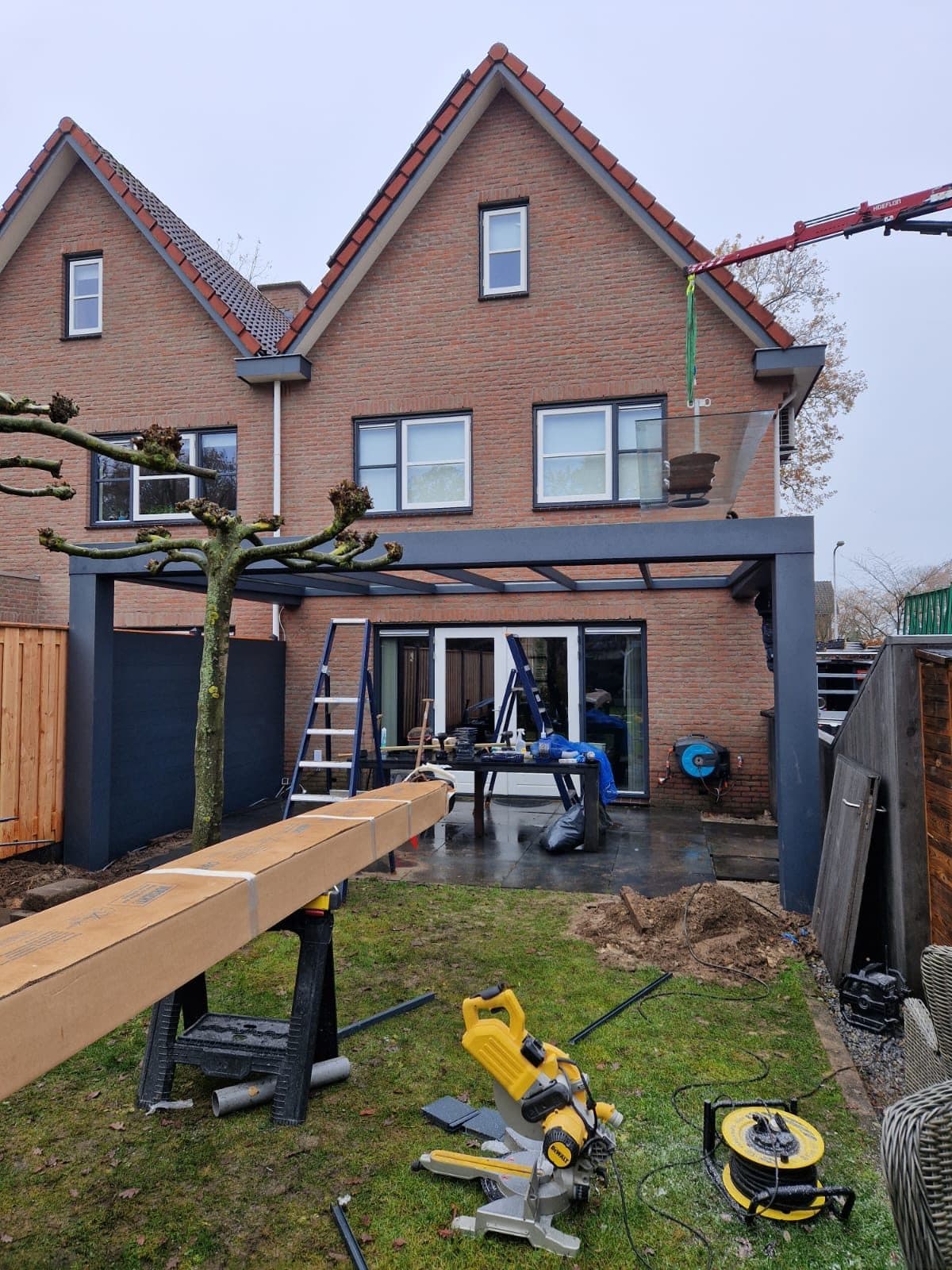 Veranda in aanbouw achter een rijwoning, met een kraan, een houten balk op schragen en gereedschap op het gras