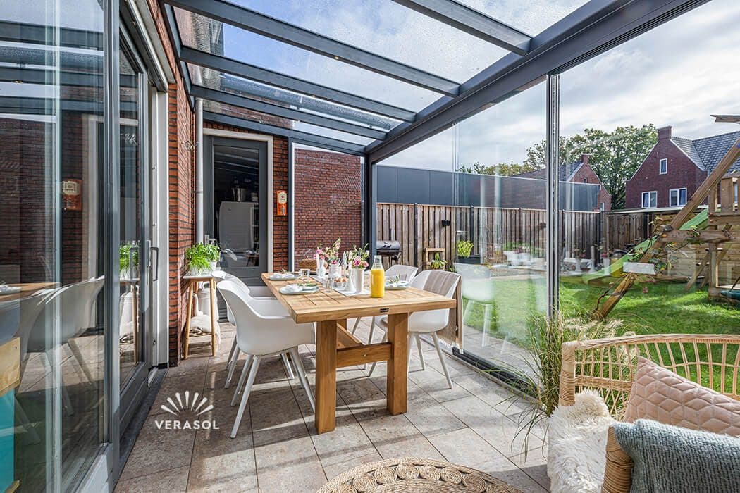 Gedekte houten tafel met witte stoelen in een antraciet tuinkamer met glazen wanden, met een speeltoestel in de tuin