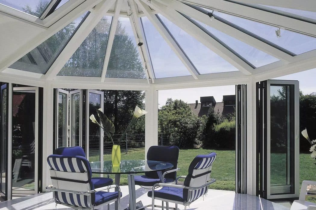 Interieur van een serre met wit glasdak en geopende vouwwand naar de tuin, met een tuinset