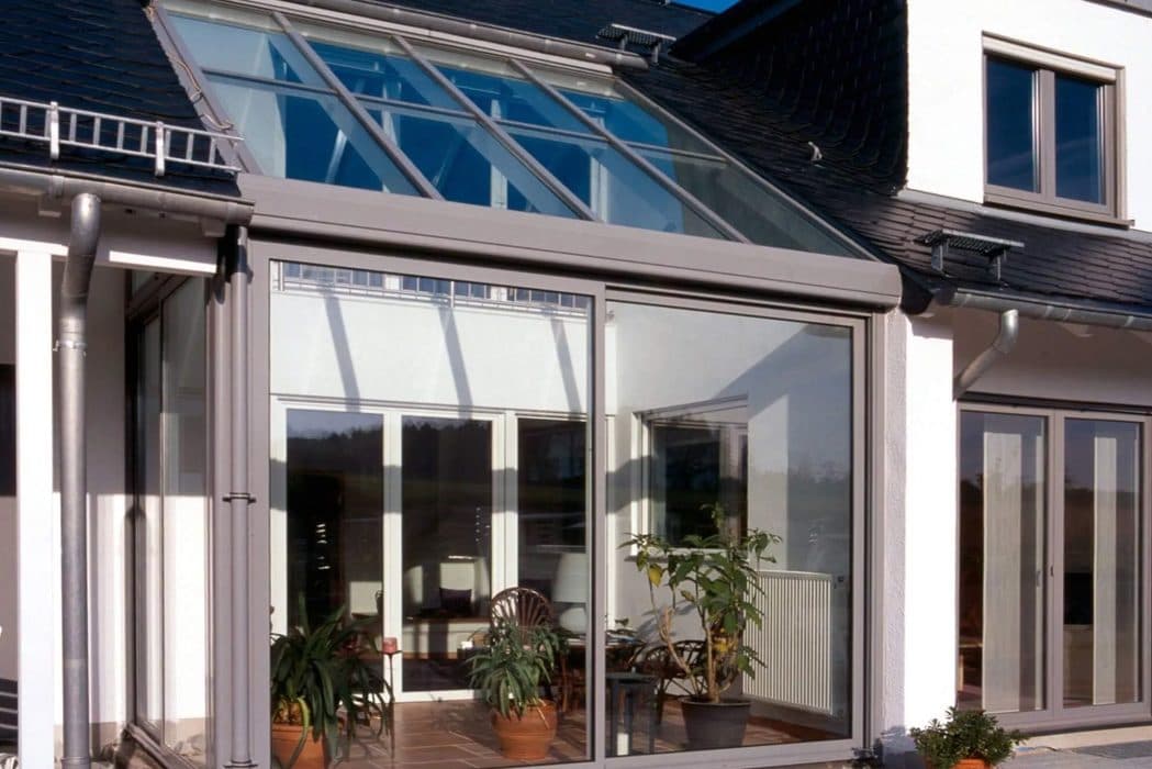 Lichtgrijze serre met glasdak en schuifpuien tegen een witte woning, met planten binnen