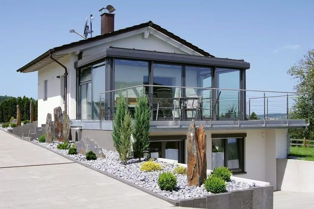 Antraciet glazen serre op het terras van een witte woning, met glazen balustrade en grindtuin
