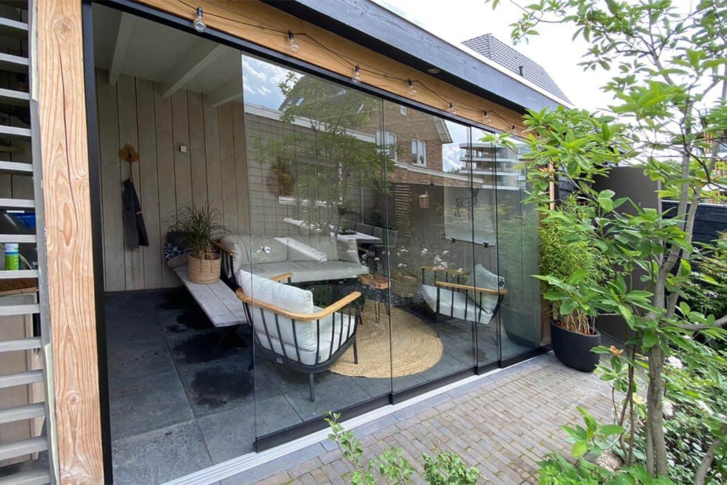 Houten overkapping met glazen schuifwanden, loungestoelen en een lichtsnoer, naast een klinkerpad