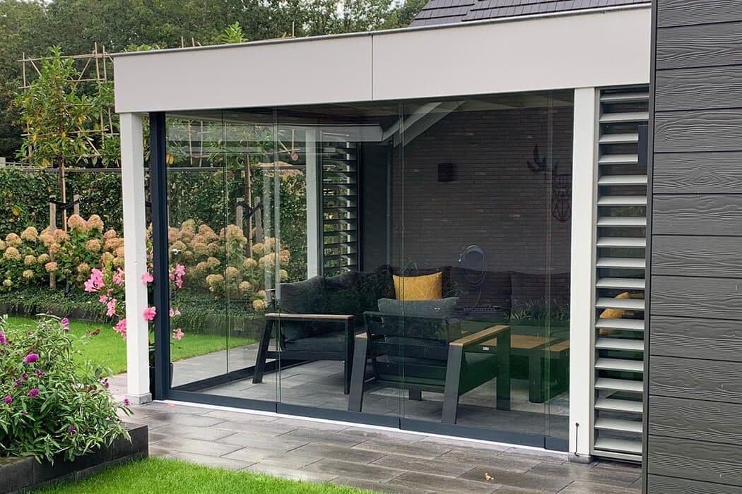 Overkapping met witte dakrand, glazen schuifwanden en een lamellenwand aan de zijkant, met loungeset in een bloemrijke tuin