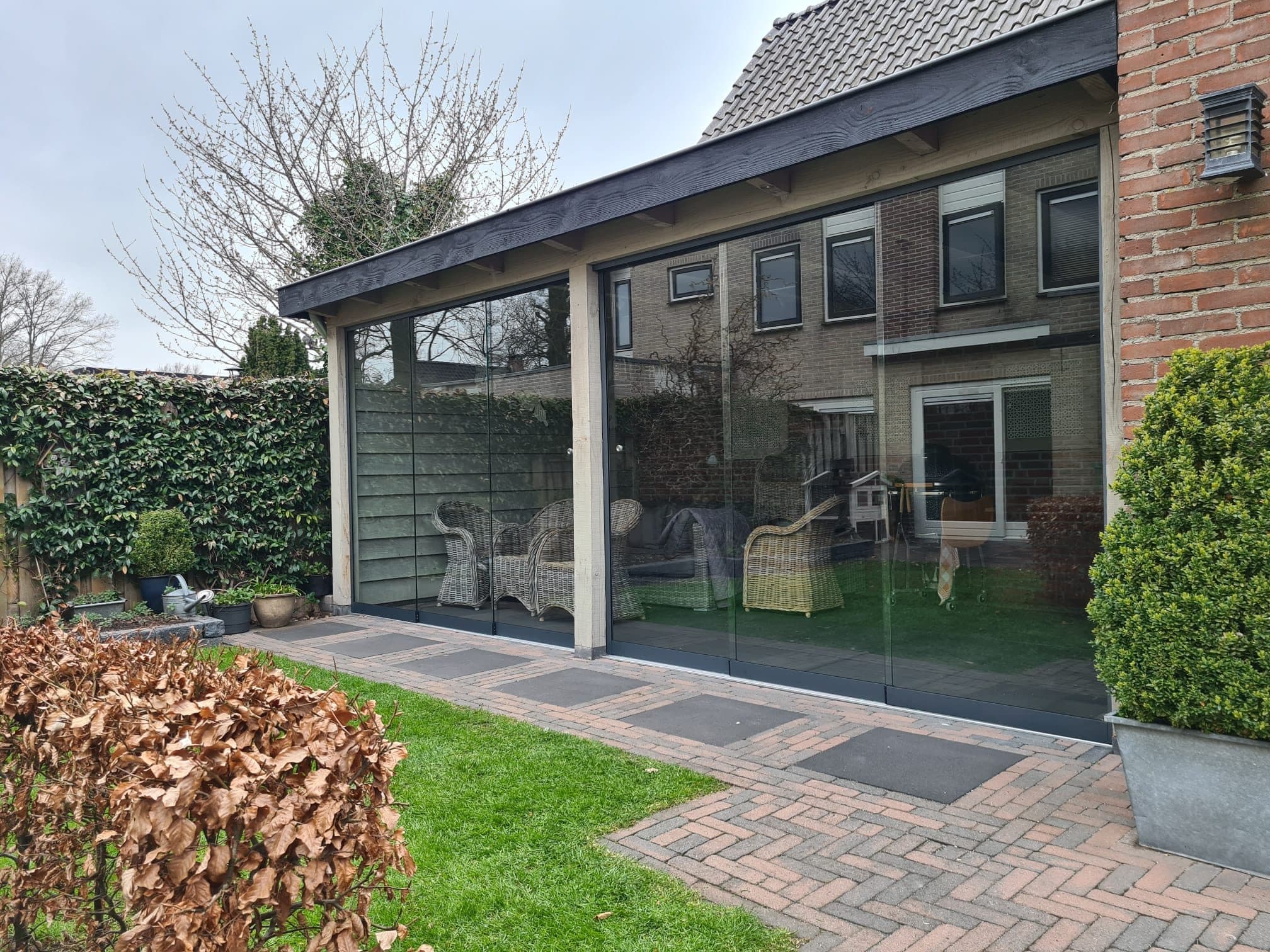 Donkere houten overkapping met glazen schuifwanden en rieten stoelen, achter in een tuin met een klinkerpad