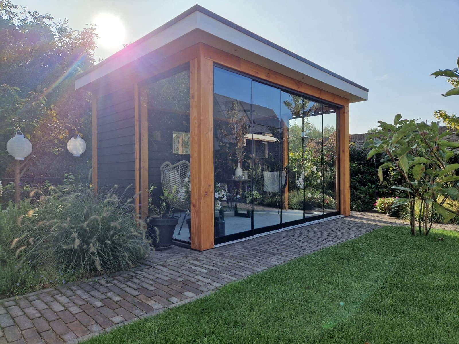Houten overkapping met glazen schuifwanden in de hoek van een tuin, met siergrassen en een klinkerpad