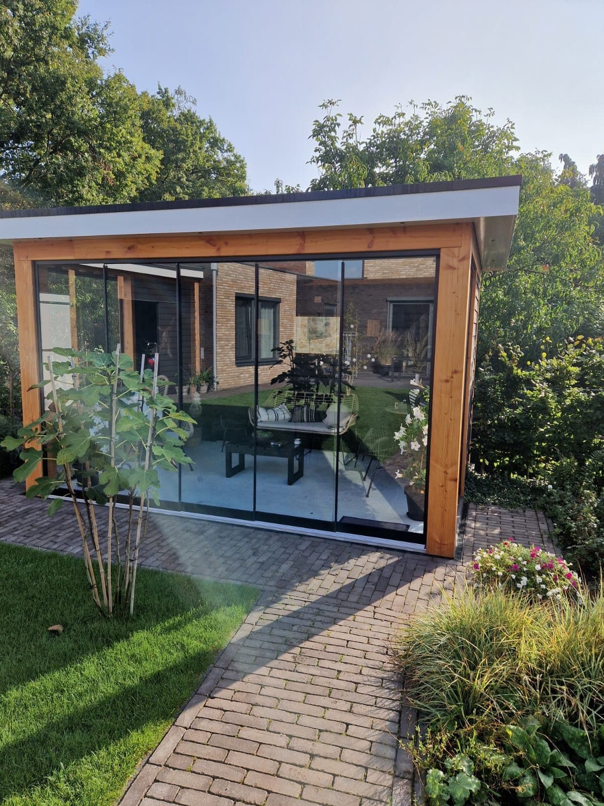 Houten overkapping met glazen schuifwanden aan het eind van een klinkerpad, met een eettafel binnen