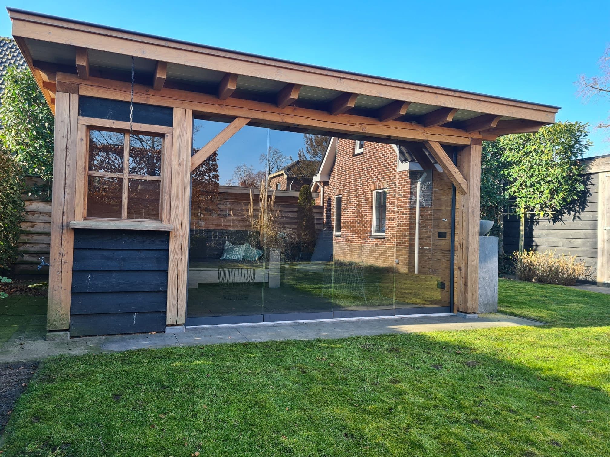 Houten overkapping met een dicht deel met raam en glazen schuifwanden, op een zonnig gazon