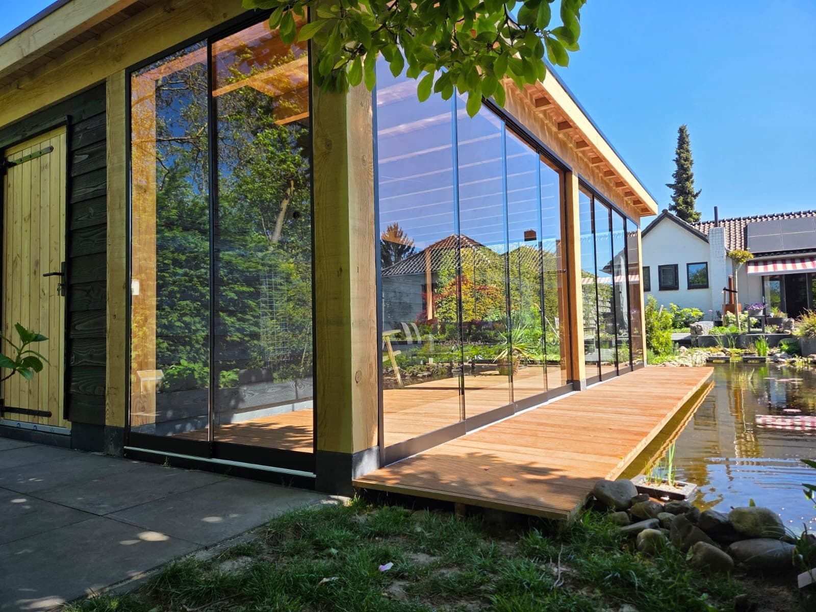 Houten overkapping met glazen schuifwanden en een vlonder langs een vijver, in de zon