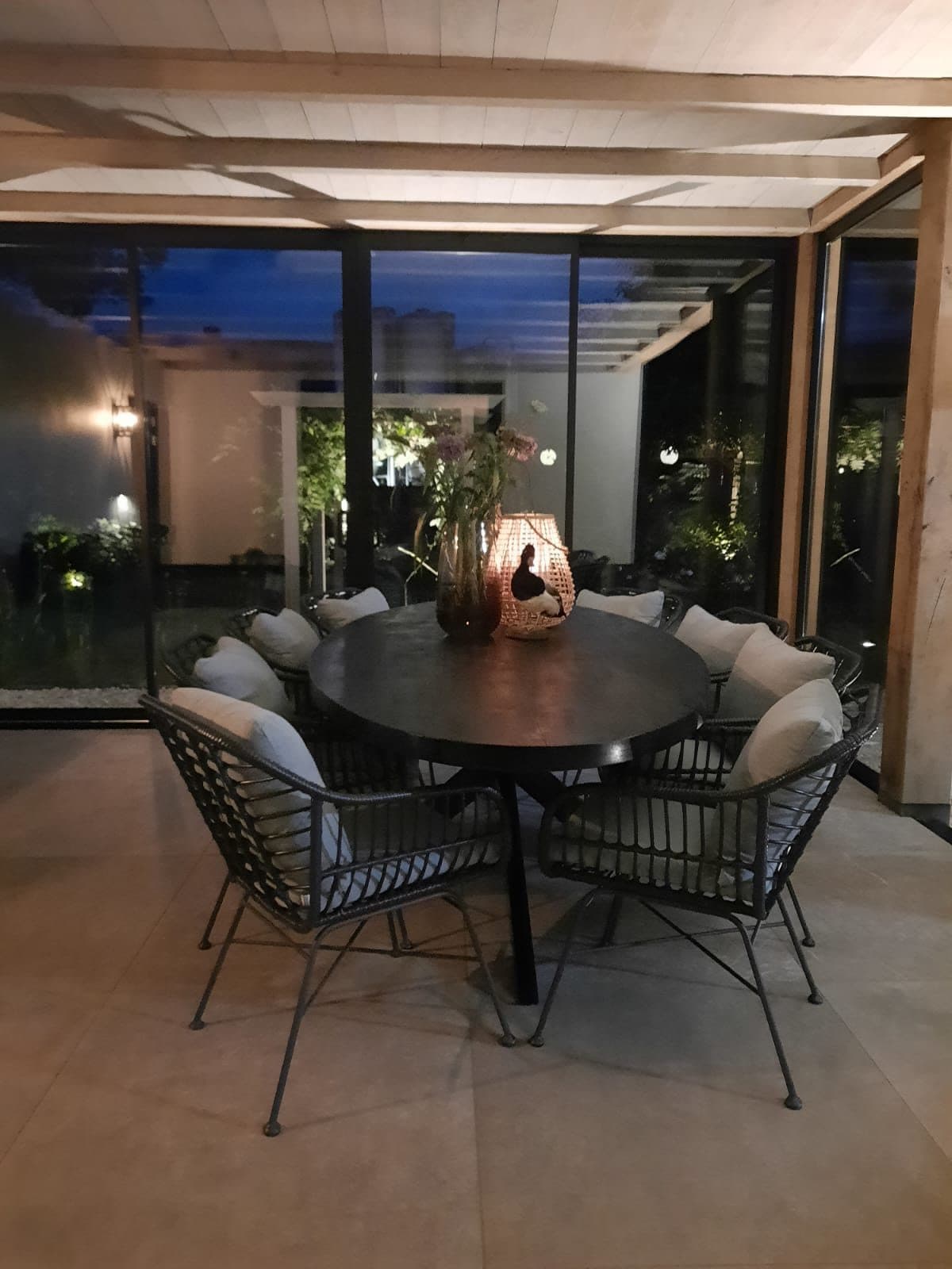 Ronde eettafel met stoelen en een lamp onder een houten overkapping met glazen wanden, 's avonds