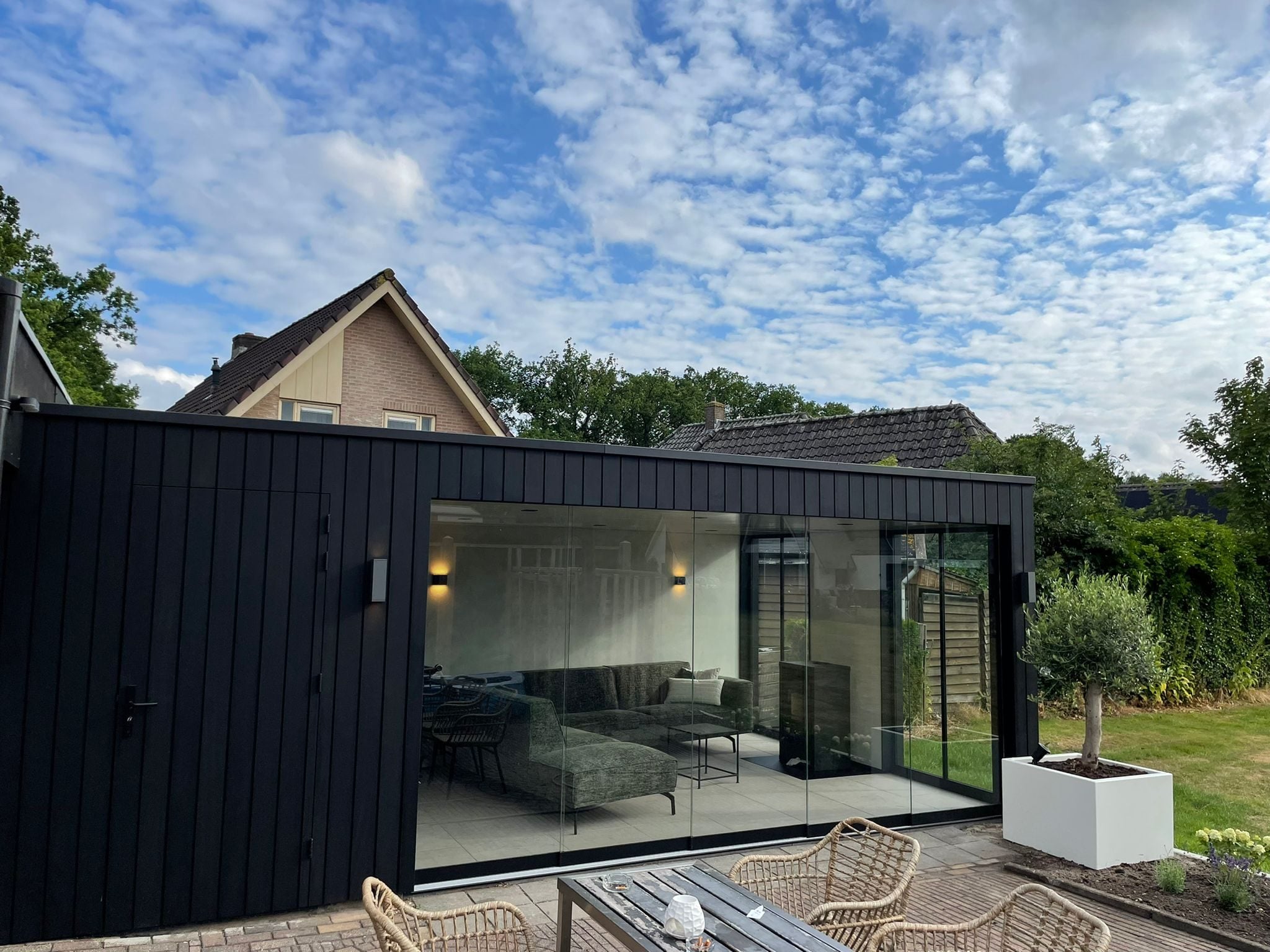 Tuinkamer met zwarte houten gevelbekleding en een glazen pui, met een zithoek binnen en rieten stoelen op het terras