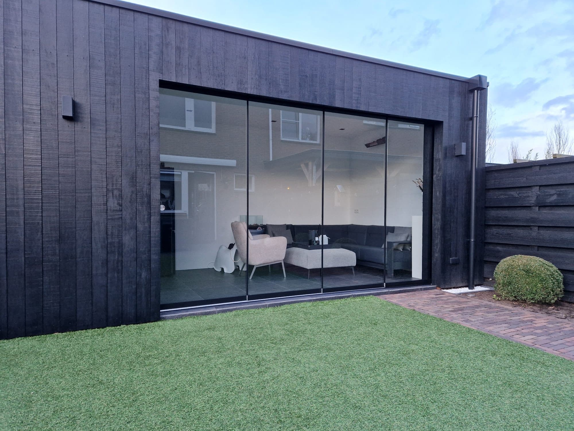 Zwarte houten tuinkamer met een brede glazen schuifwand, met een zithoek binnen en kunstgras ervoor