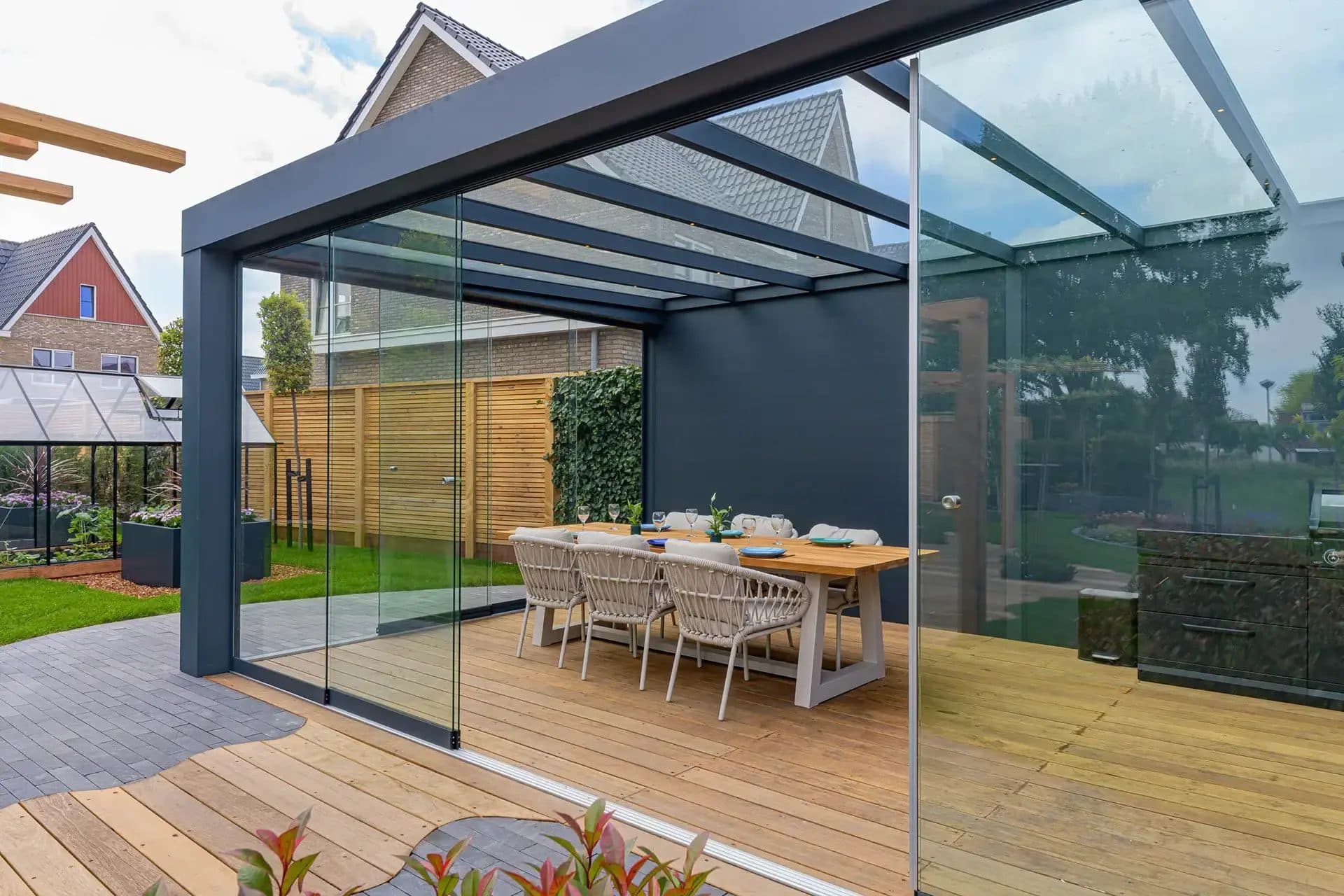 Vrijstaande antraciet tuinkamer met glasdak en glazen schuifwanden op een houten vlonder, met gedekte eettafel en een kas op de achtergrond