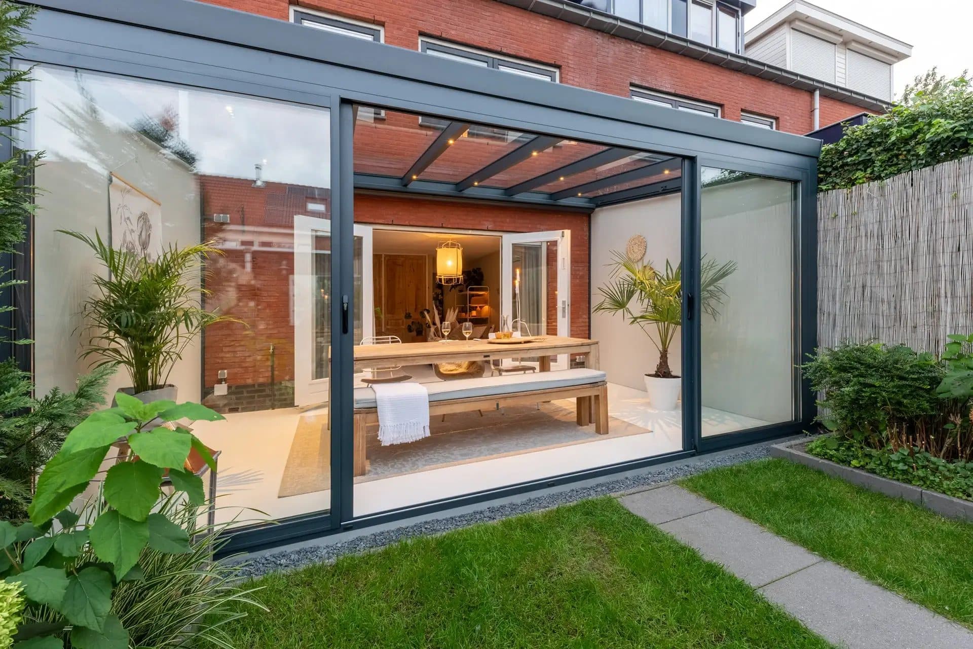 Antraciet tuinkamer met glasdak en aluminium schuifpui achter een rijwoning, met gedekte eettafel binnen