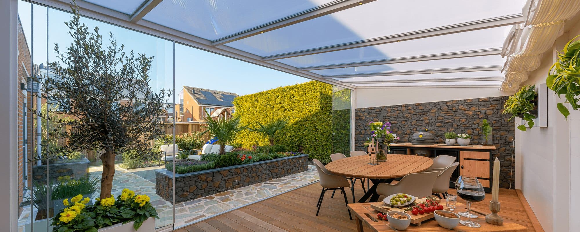 Eettafel met gekleurd servies in een tuinkamer met glasdak en glazen wanden op een houten vloer, met zicht op de tuin