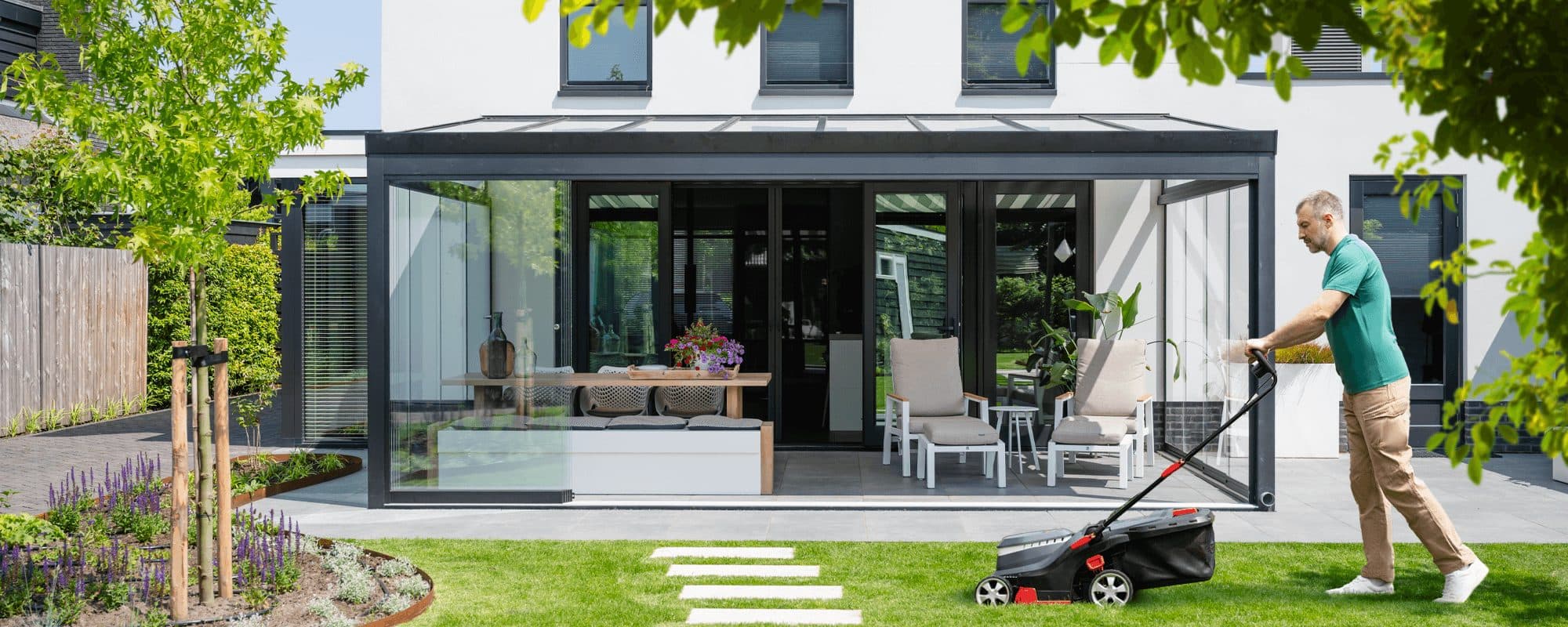 Antraciet tuinkamer met glazen wanden aan een witte woning, met een man die het gazon maait