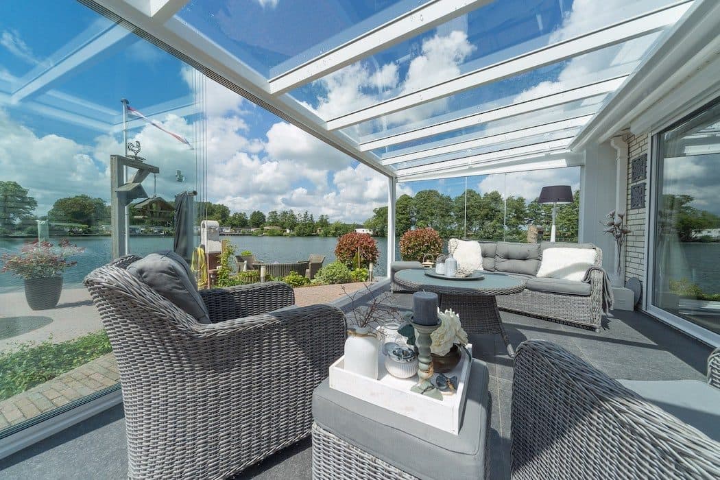 Interieur van een witte veranda met glasdak en glazen wanden, met rieten loungestoelen en uitzicht op het water