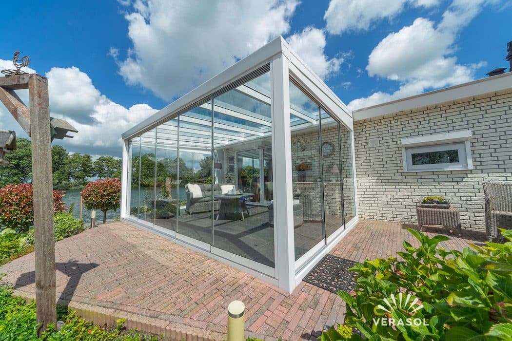 Witte veranda met glasdak en rondom glazen wanden op een klinkerterras naast een witte bakstenen woning