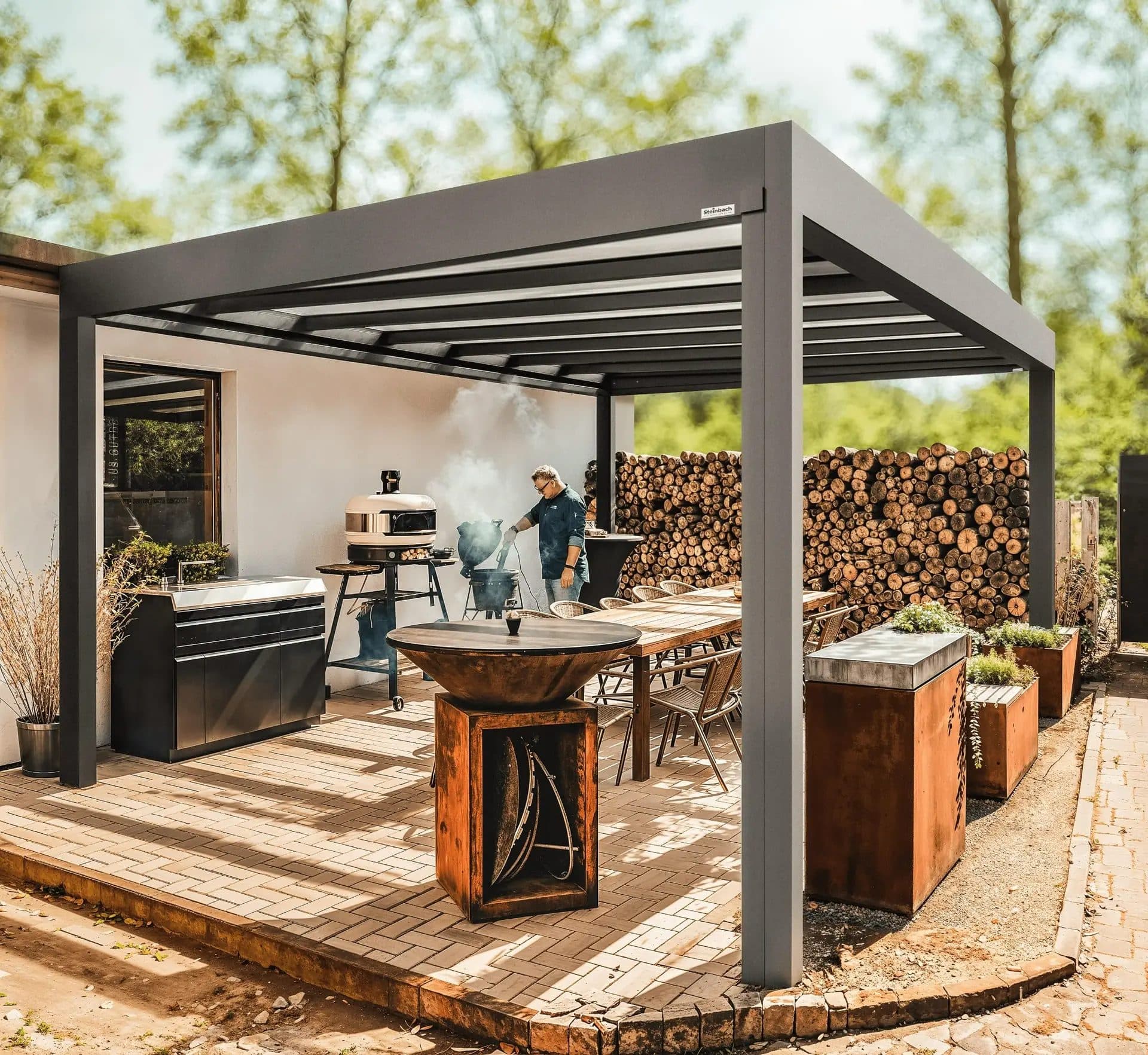 Open grijze overkapping met vlak dak boven een buitenkeuken met barbecue, houtopslag en een lange tafel