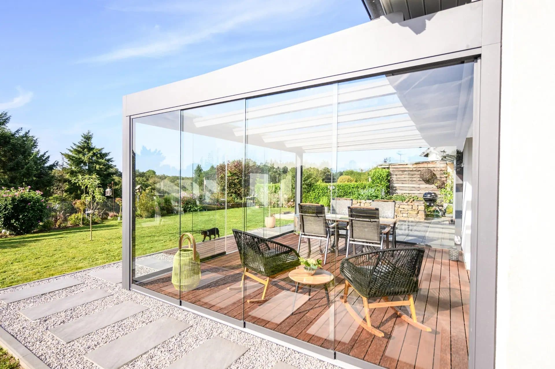 Lichtgrijze veranda met vlak ogend glasdak en glazen schuifwanden aan een witte woning, met tuinstoelen op een houten vlonder