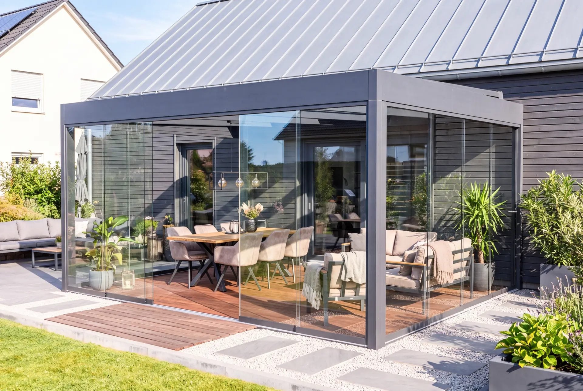 Grijze kubusvormige veranda met rondom glazen schuifwanden tegen een woning met donkere houten gevel, met eettafel en loungeset op een houten vlonder