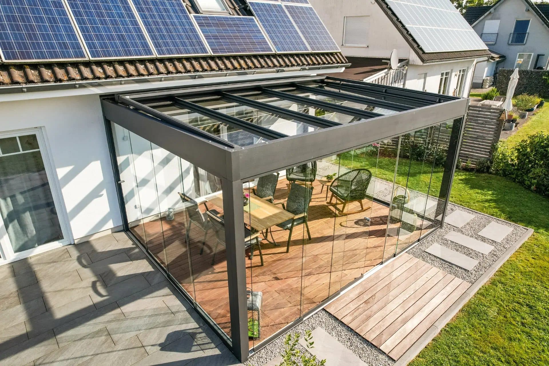 Antraciet veranda met glasdak en glazen wanden tegen een witte woning met zonnepanelen op het dak, van bovenaf gezien