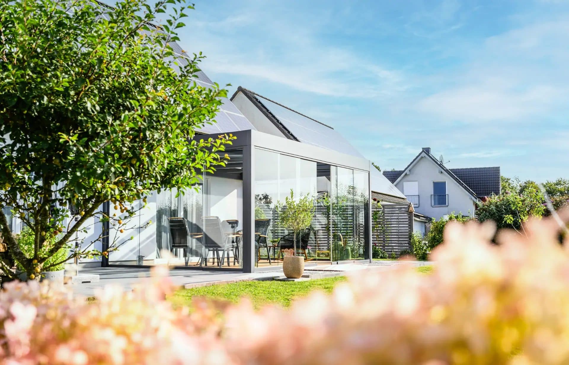 Veranda met glazen wanden tegen een witte woning met puntdak, gezien vanuit de tuin met bloeiende struiken op de voorgrond