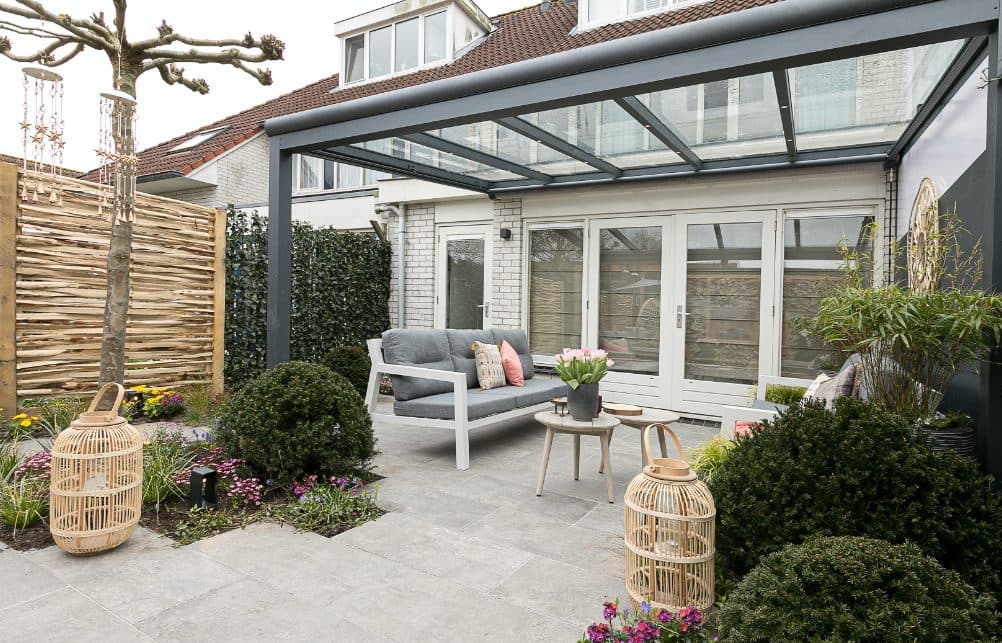 Antraciet veranda met glasdak achter een rijwoning, met loungebank, lantaarns en borders in een stadstuin