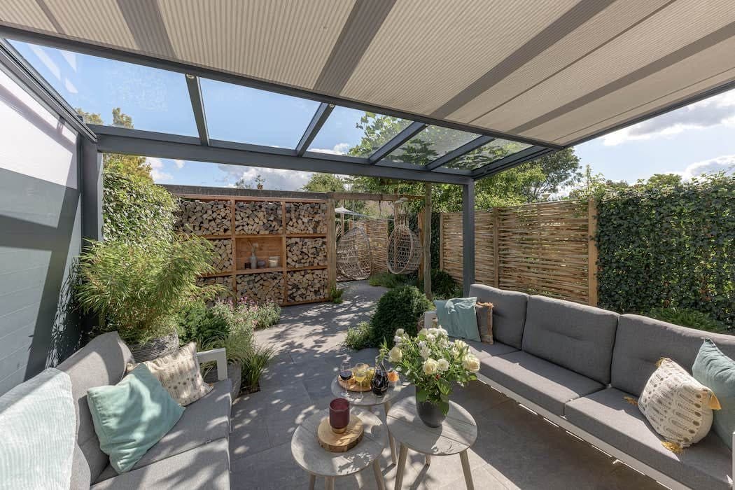 Hoekbank en bijzettafels onder een veranda met glasdak en uitgerold doek onder het glas, in een tuin met wilgenschermen
