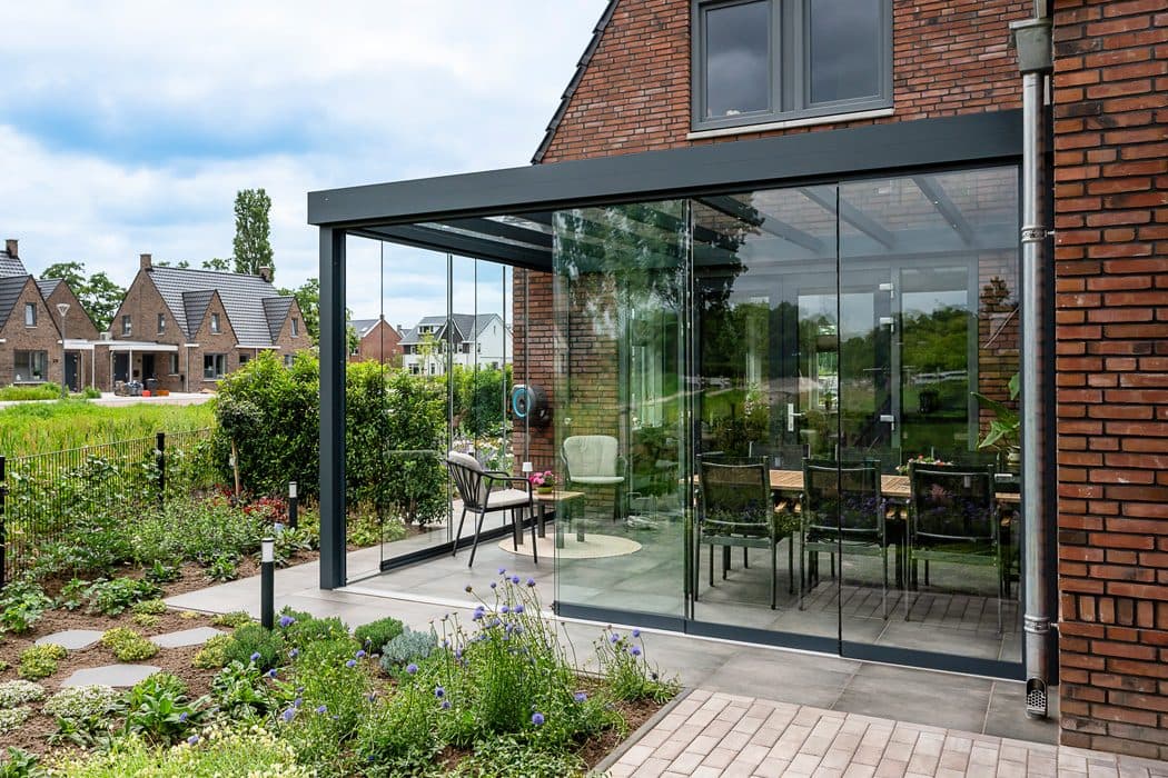 Antraciet veranda met strak, vlak dak en rondom glazen wanden tegen een bakstenen woning, gezien vanuit een tuin met borders