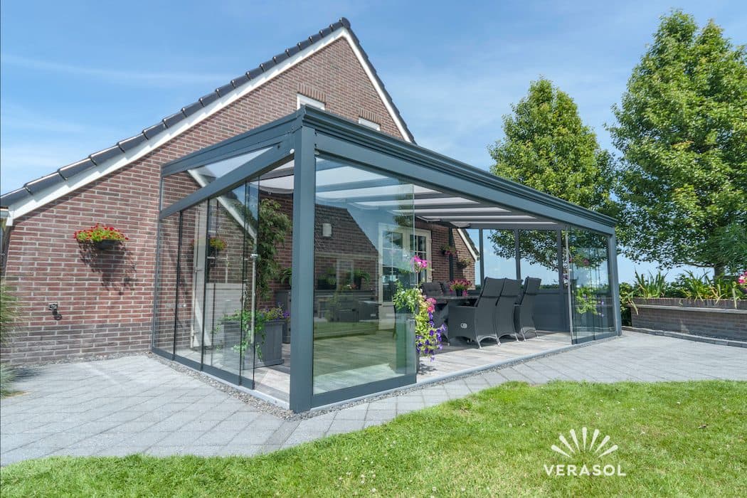 Antraciet veranda met zichtbare dakhelling en glazen wanden tegen een bakstenen woning met puntdak, gezien vanaf het gazon