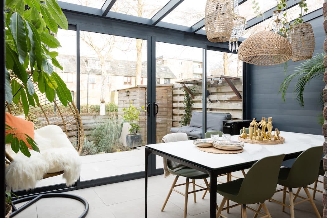 Eettafel met groene stoelen en rieten hanglampen onder een antraciet veranda met glazen schuifwanden