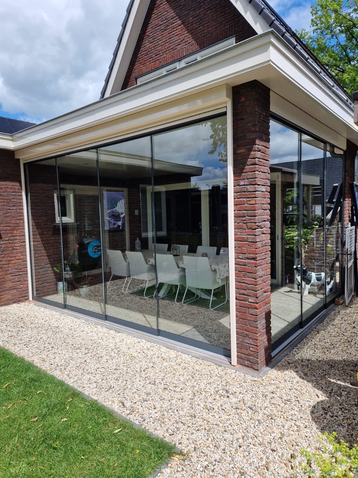 Glazen schuifwanden onder de witte overstek van een bakstenen woning, met een lange eettafel erachter