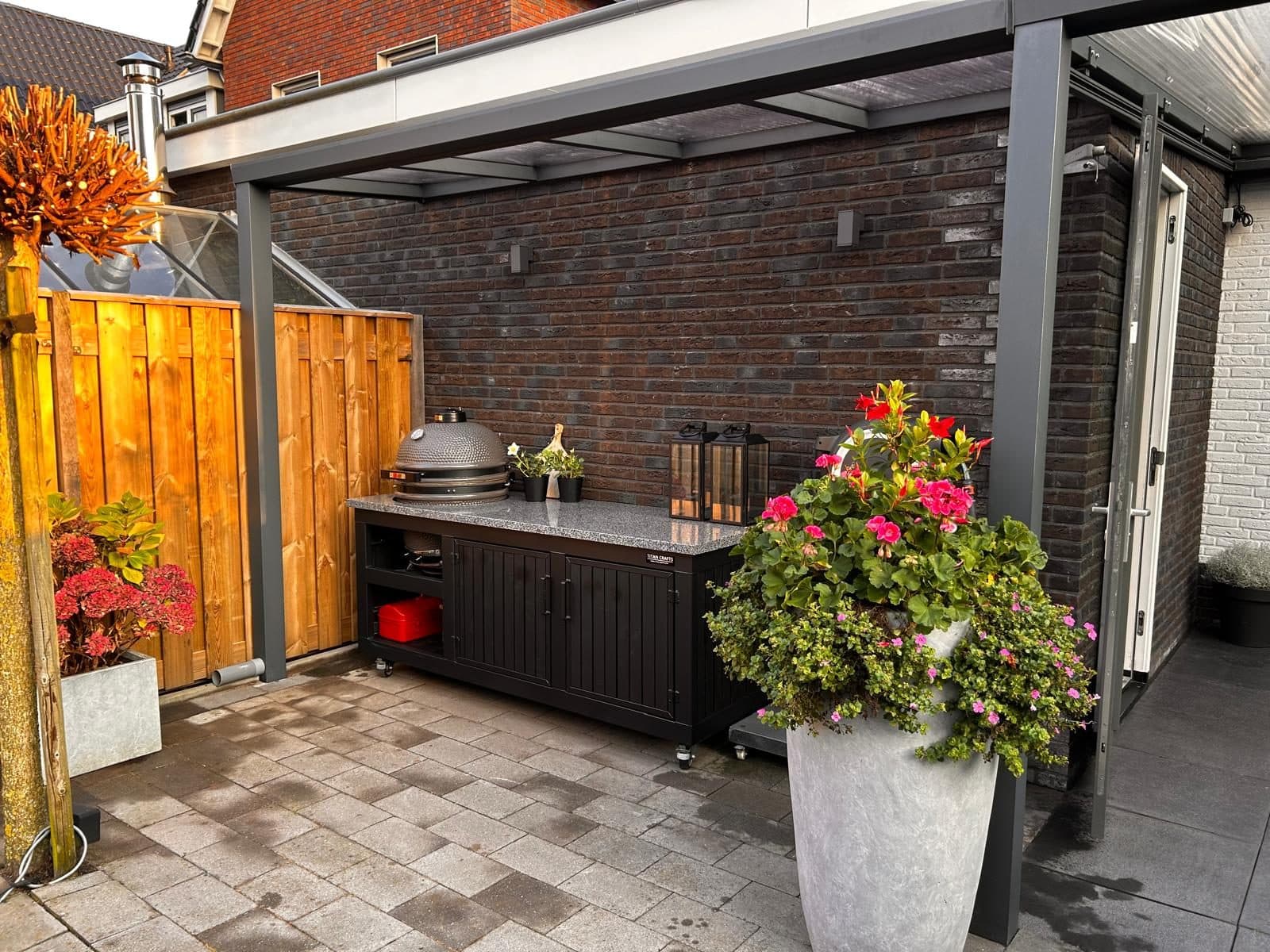 Buitenkeuken met barbecue onder een antraciet overkapping, met een grote pot bloemen ervoor