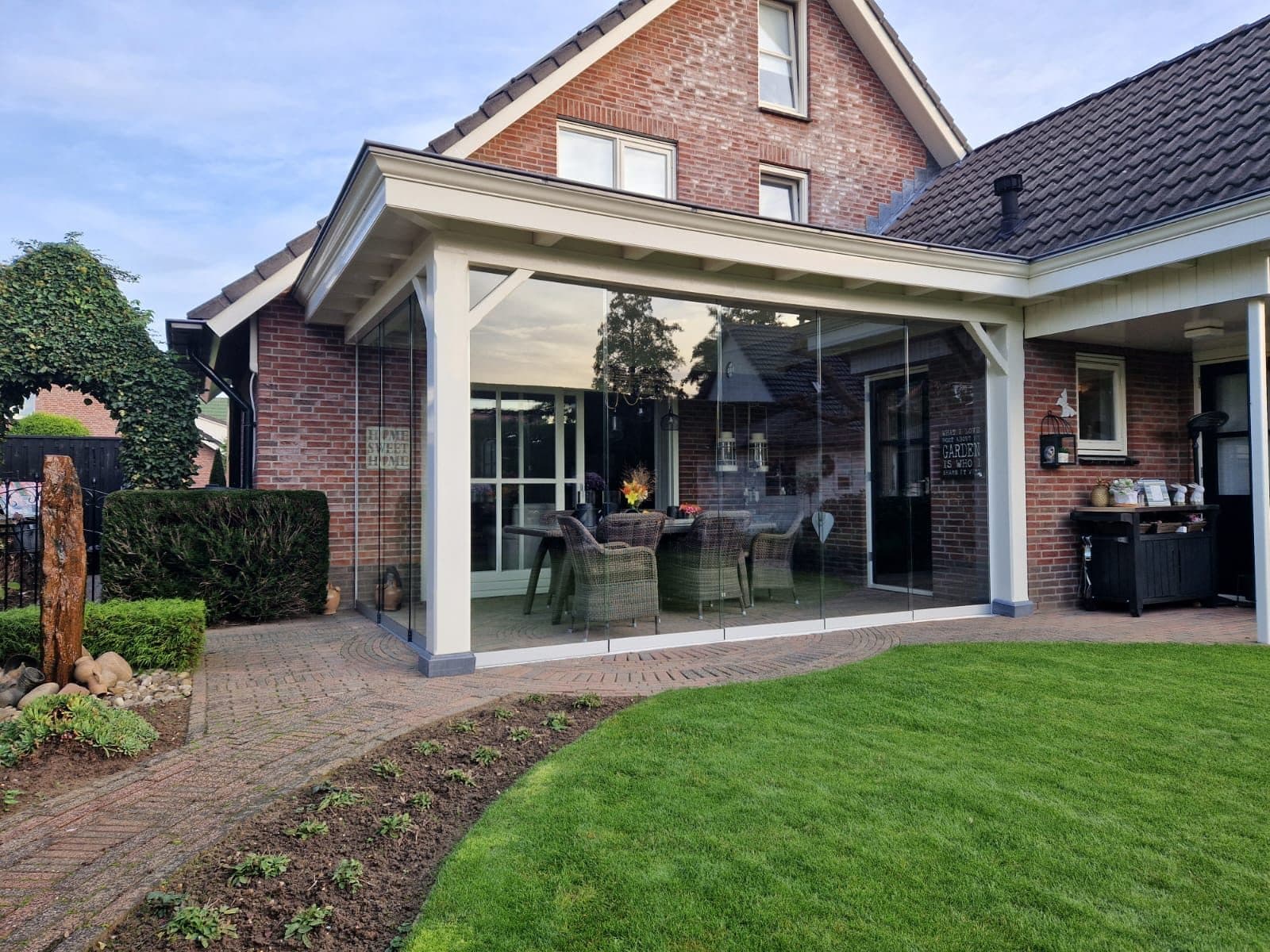 Crèmekleurige overkapping met glazen schuifwanden aan een bakstenen woning, met een gebogen gazon ervoor