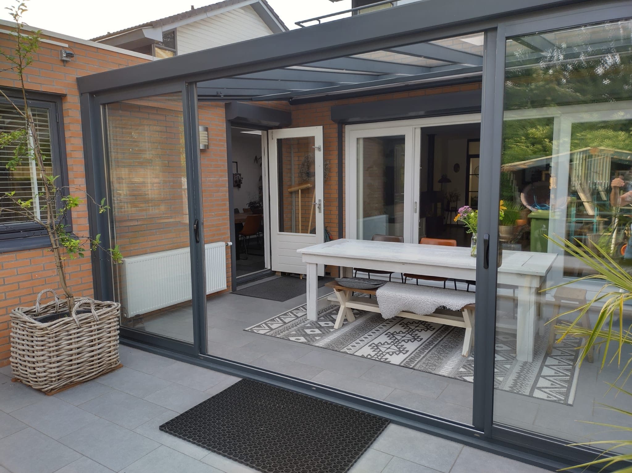 Antraciet veranda met glazen schuifwanden over een witte eettafel met bank, aan een bakstenen woning