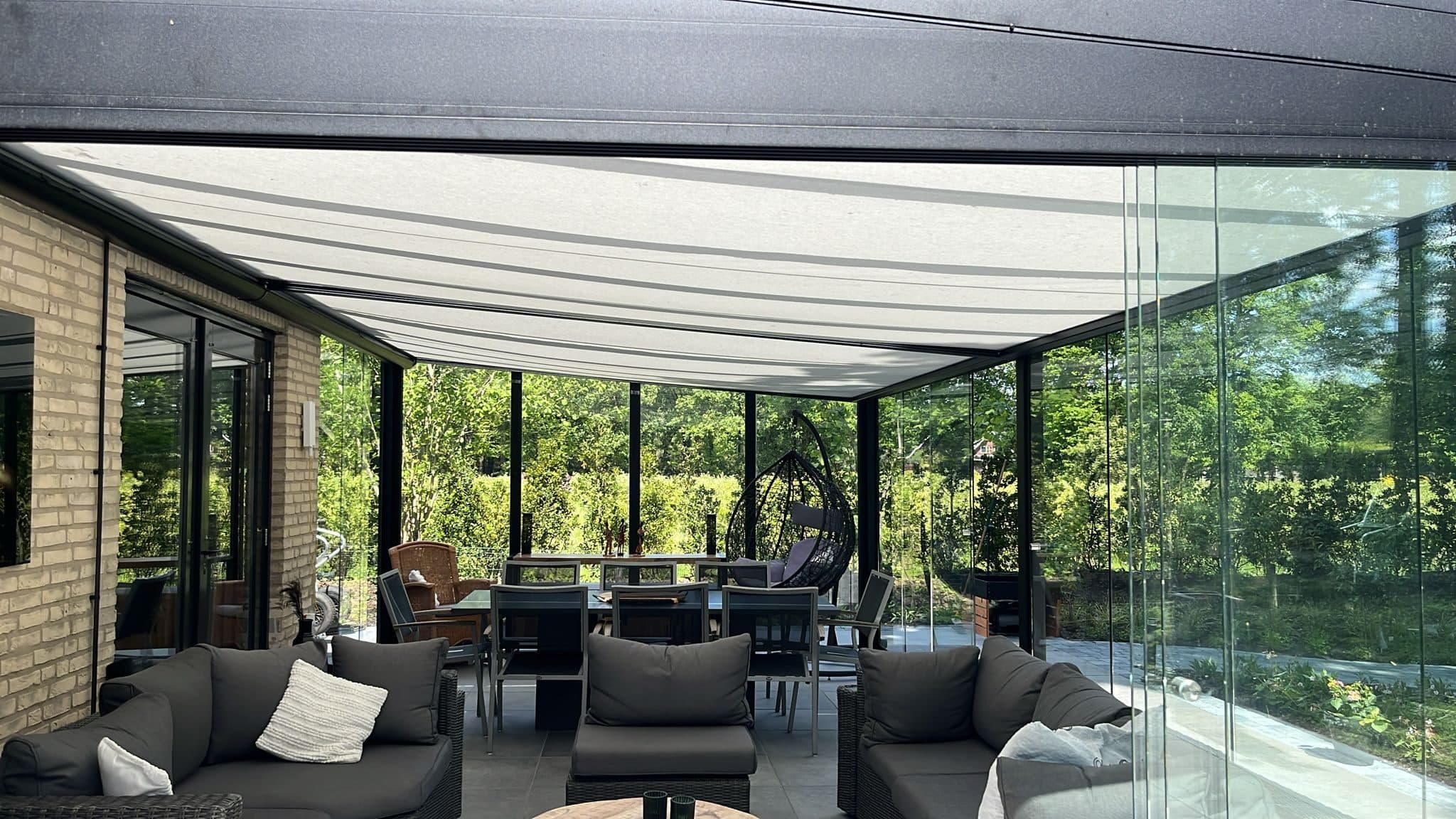 Loungebanken en een eettafel onder een veranda met wit doek onder het dak en glazen wanden rondom, met zicht op het groen