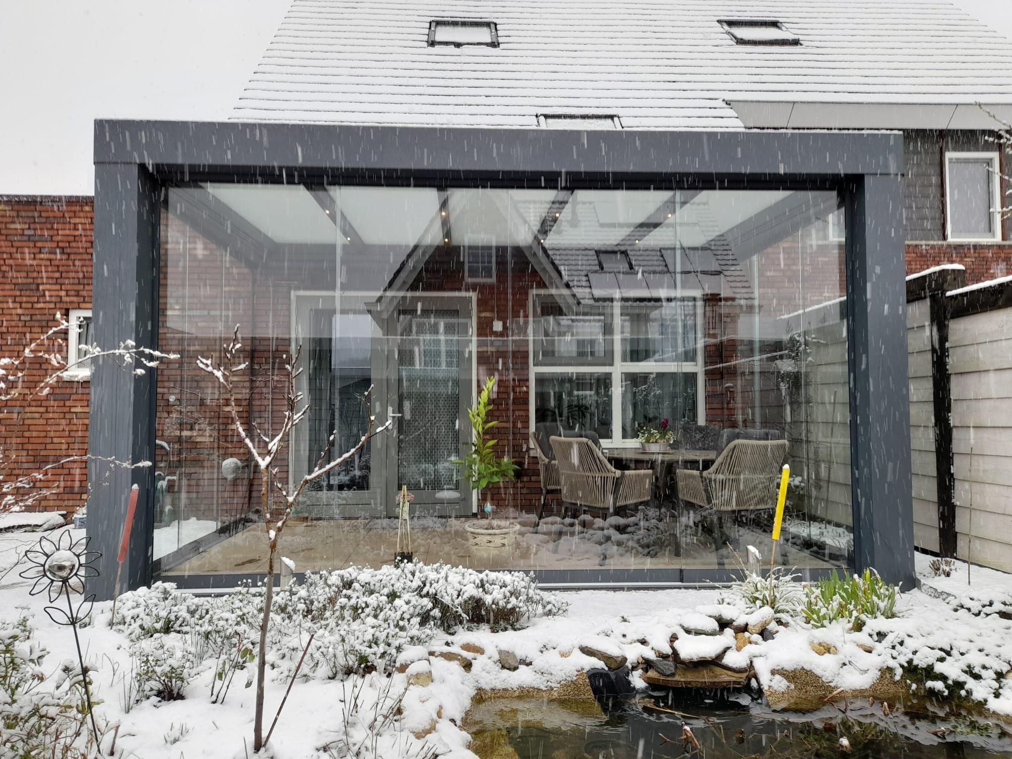 Antraciet veranda met glazen schuifwanden en een eethoek, in een tuin met sneeuw en een vijver