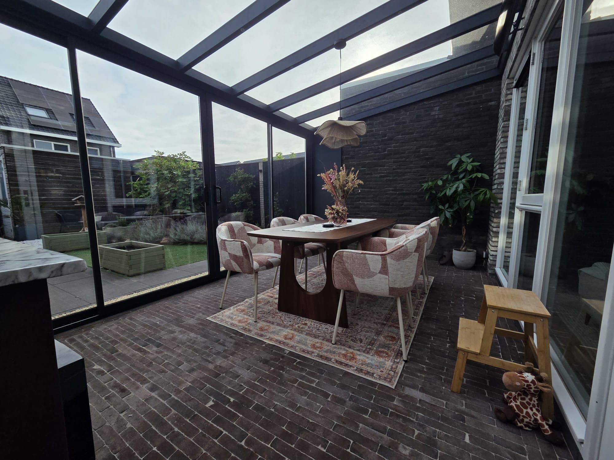 Eettafel met roze stoelen onder een antraciet glasdak, met glazen schuifwanden aan de tuinkant