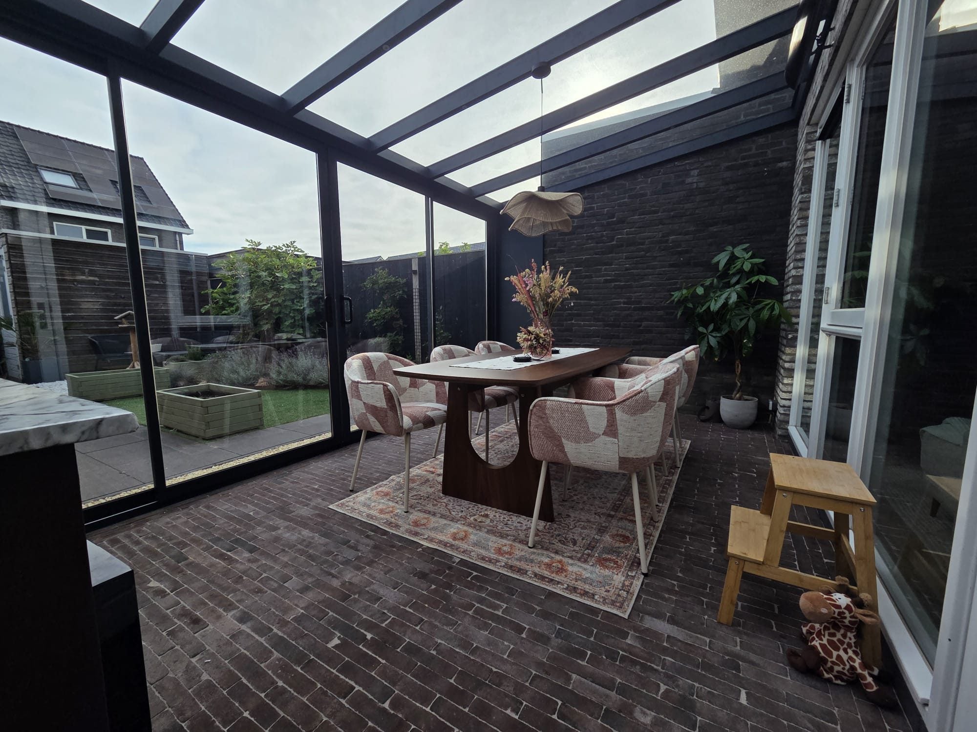 Eettafel met roze stoelen onder een antraciet glasdak, met glazen schuifwanden aan de tuinkant